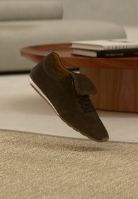 Σκούρο καφέ σουέτ αθλητικό παπούτσι με μαύρα κορδόνια και διακοσμητικό καπακι, αιωρούμενο πάνω από μπεζ υφαντό χαλί σε σαλόνι με ξύλινο τραπέζι και βιβλία.