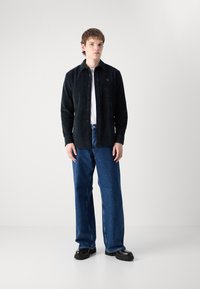 Camicia in velluto a coste nera con bottoni sopra una maglietta bianca, abbinata a jeans larghi in denim blu e scarpe chunky nere. Design semplice con cuciture visibili.