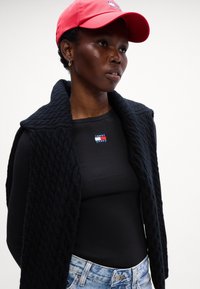 Gilet nero lavorato a maglia con colletto alto sopra una maglietta nera aderente a maniche lunghe con un piccolo logo rosso, bianco e blu. Cappellino rosso indossato di lato.