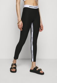 Svarta leggings med en vit märkesrand ned längs sidan. Sval och smidig textur, åtsittande design, kombinerade med svarta sandaler med gulddetaljer.