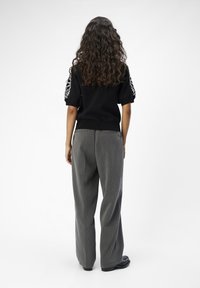 Mujer con cabello largo y rizado, vistiendo una blusa negra de manga corta con mangas blancas con estampado floral y pantalones grises sueltos, de pie de espaldas.