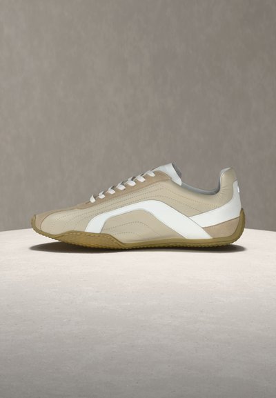 Baskets basses beige clair et blanches avec semelle en caoutchouc beige et lacets blancs, présentées sur une surface en pierre claire sur un fond gris.