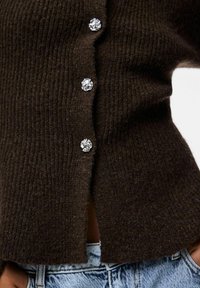 Brūns ribota adīva kardigāns ar pieciem apaļiem, caurspīdīgiem kristālu pogām un sānu šķēlēm. Kombinēts ar gaiši ziliem džinsu auduma biksēm. Mīksta tekstūra.