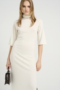 Krēmkrāsas turtleneck kleita ar trīs ceturtdaļu garuma piedurknēm, gluda tekstūra, pieguloša forma un sānu aukla, kopā ar tumšu ādas rokassomu.