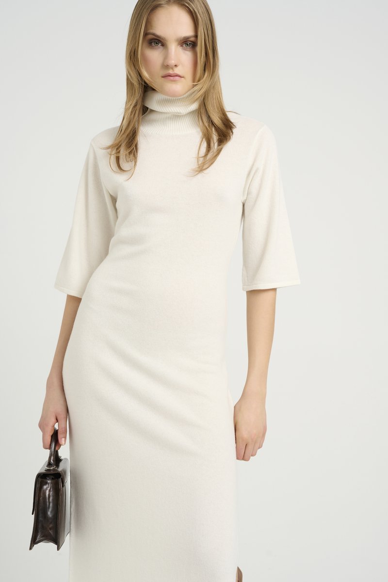 Krēmkrāsas turtleneck kleita ar trīs ceturtdaļu garuma piedurknēm, gluda tekstūra, pieguloša forma un sānu aukla, kopā ar tumšu ādas rokassomu.