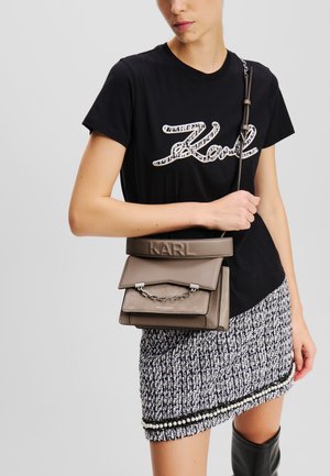 Tan leren handtas met drie gestapelde compartimenten, zilveren kettingaccent, logo in reliëf aan de voorkant en verstelbare schouderband. Zwarte t-shirt met wit logo en gestructureerde zwart-witte rok met parelaccenten.