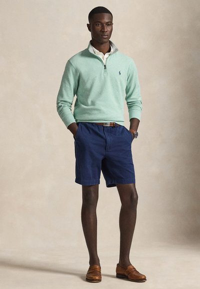 Camisola verde clara com fecho de correr sobre uma camisa branca com colarinho, calções azul-marinho, cinto castanho e sapatos castanhos sem atacadores. Design simples, estilo casual.