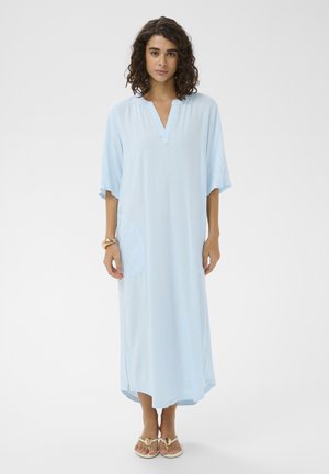 Femme aux cheveux bouclés bruns, portant une longue robe bleue claire avec des poches et des sandales beige, debout devant un fond blanc uni.