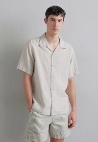 Camicia in lino beige chiaro a maniche corte con colletto a campana, chiusura frontale con bottoni, abbinata a pantaloni corti a righe chiare. Tessuto morbido, vestibilità comoda.