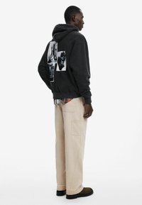 Felpa nera con grafiche stampate sul retro, abbinata a pantaloni cargo beige a gamba larga e scarpe scure. Tessuti morbidi e casual.