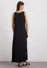 Robe maxi sans manches noire avec un col rond et des fentes latérales, fabriquée dans un matériau lisse et léger. Les sandales ont un accent en boucle.