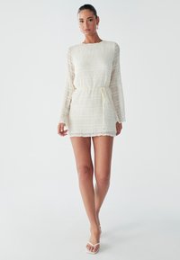 Weißes Mini-Kleid mit langen Ärmeln, strukturierter Stoff und Taillengürtel. Mit Rundhalsausschnitt und gewelltem Saum. Kombiniert mit weißen Sandalen.