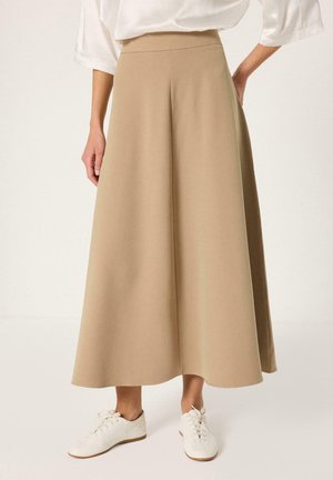 Γυναίκα που φοράει μπεζ μίντι φούστα με φαρδιά φούστα και λευκά αθλητικά παπούτσια, στέκεται με το ένα χέρι στη μέση μπροστά σε απλό φόντο.