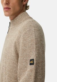 Man in een beige gehaakte trui met een ritskraag en een zwart-geel logo op de bovenarm.