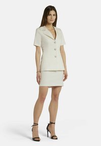 Blazer en lin blanc à manches courtes, col à revers et trois boutons, associé à une mini-jupe blanche ajustée et des escarpins noirs à brides.