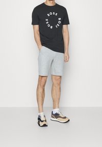 Man som bär en svart T-shirt med cirkulär text "BOSS", ljusgrå shorts, vita strumpor och beige, marinblå och bruna sneakers, står med ena handen i fickan.