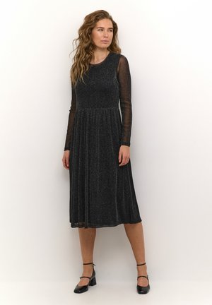 Black long-sleeve dress with a fitted bodice, flared skirt, and shimmer texture. Paired with black block-heel shoes and ankle straps.
