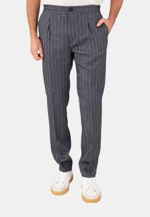 Uomo che indossa pantaloni grigi a righe con pinces e sneakers bianche, in piedi con una mano in tasca.