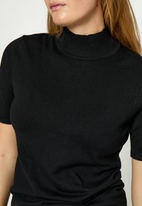 Woman wearing a black short-sleeve knit top with a ribbed mock neck, shown from shoulders to waist against a plain background.