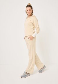 Felpa beige tagliata con fessure laterali e logo arancione, abbinata a pantaloni larghi beige coordinati. Sneakers grigie completano l'outfit.
