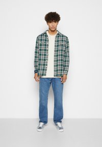 Groen en marineblauwe geruite flanellen shirt over een crèmekleurig top, gecombineerd met lichtblauwe jeans en gestreepte sneakers, staande tegen een witte achtergrond.
