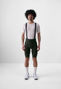 Witte mesh korte mouwen shirt, groene gewatteerde fietsshorts met zwarte bretels, en witte sokken met fietsschoenen, staand op een effen achtergrond.