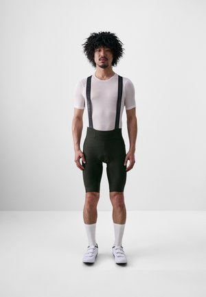 Mesh white short-sleeve shirt, green padded cycling shorts with black suspenders, and white socks with cycling shoes, standing on a plain background.