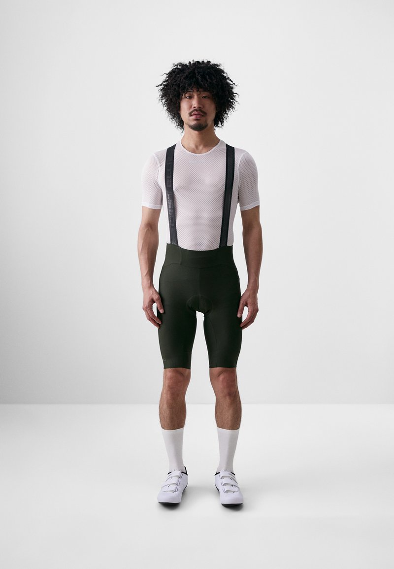 Witte mesh korte mouwen shirt, groene gewatteerde fietsshorts met zwarte bretels, en witte sokken met fietsschoenen, staand op een effen achtergrond.