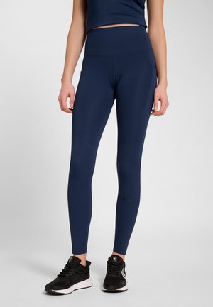 Frau trägt marineblaue Leggings mit hoher Taille und schwarze Sportschuhe, steht vor einem schlichten hellgrauen Hintergrund.