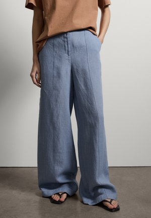 Persona che indossa pantaloni larghi in lino azzurro chiaro, top marrone a maniche corte e sandali infradito neri, in piedi su un pavimento grigio contro una parete bianca.