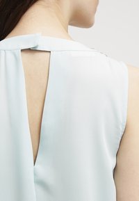 Blusa senza maniche di colore blu chiaro con un'apertura a forma di V sulla schiena, leggero lucido e chiusura al collo sicura con un piccolo anello di tessuto.