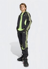 Teenager steht selbstbewusst in einem schwarzen und neon-grünen Adidas Trainingsanzug mit passender Jacke, Hose und schwarzen Sneakers.
