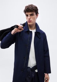 Νέος άνδρας με κοντά σγουρά μαλλιά, φοράει μπλε σκούρο παλτό πάνω από άσπρο πουκάμισο, κρατώντας μια μαύρη τσάντα στον ώμο του, με φόντο ένα απλό λευκό φόντο.