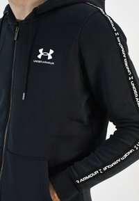 Sudadera con cremallera negra y un bolsillo frontal, presenta un logo blanco de Under Armour en el pecho y cinta con el logo a lo largo de las mangas. Textura de tela suave.