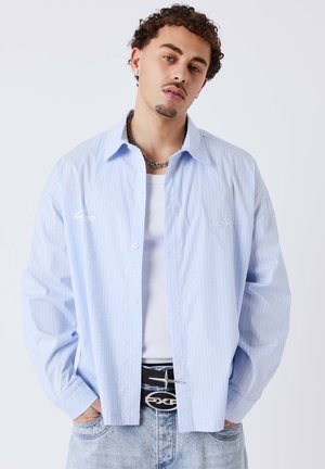 Hombre con pelo rizado que lleva una camisa de rayas azul claro sobre una camiseta blanca sin mangas, vaqueros claros, collar de cadena y cinturón negro con hebilla de estrella.
