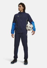 Navy sports tracksuit with blue accents, made of lightweight material. Includes a zip-up jacket and matching pants. Holding a soccer ball.