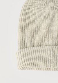 Πλεκτός σκούφος κρεμ χρώματος με διπλωμένο ρεβέρ, που δείχνει τη ριμπ υφή και λεπτομέρεια σε κοντινό πλάνο σε απλό φόντο.