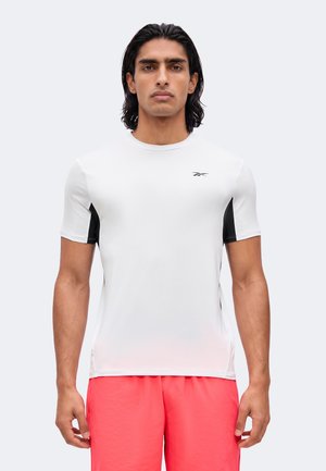 Jeune homme aux cheveux mi-longs foncés, portant un t-shirt de sport blanc et noir et un short rouge vif, se tenant face à l'avant.
