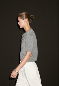 Cardigan grigio a maniche corte in maglia con tessuto testurizzato e dettagli in bottone, abbinato a pantaloni bianchi a vita alta. Sfondo semplice.