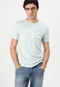 T-shirt di cotone verde chiaro con maniche corte, scollatura rotonda e una stampa grafica circolare sul petto sinistro. Indossato con jeans in denim blu.