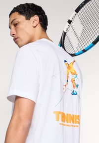 Camiseta de algodón blanca con un gráfico a color de un jugador de tenis en la espalda, con un texto grande en naranja que dice "TENNIS."