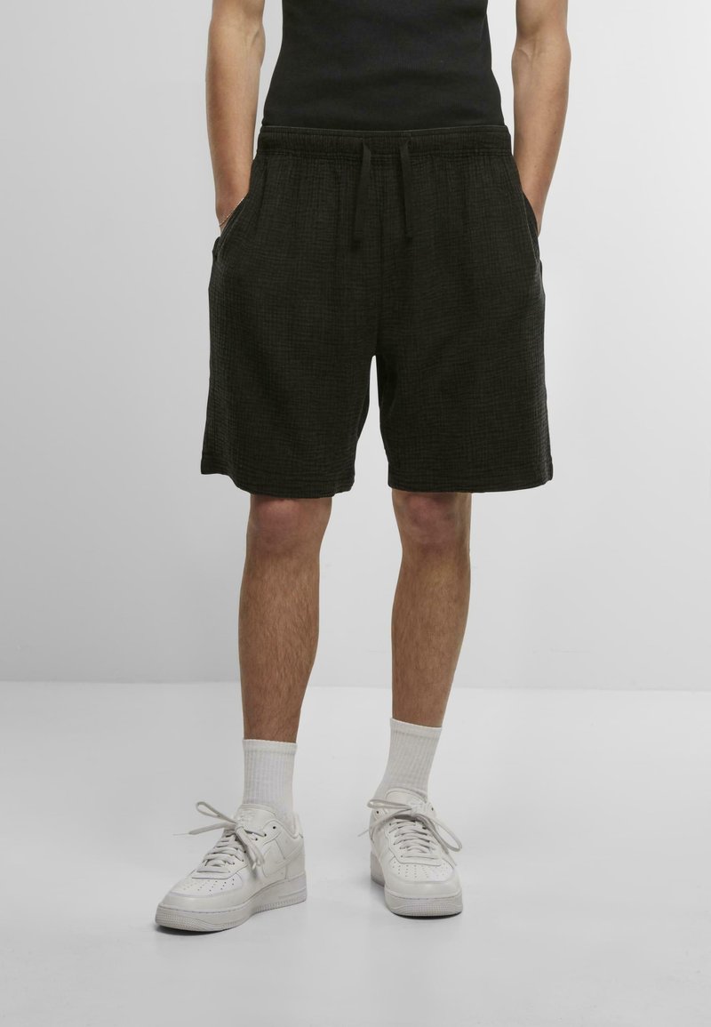 Shorts noirs en tissu texturé, dotés d'une ceinture élastique, de poches latérales et d'une coupe décontractée, assortis à des baskets blanches et des chaussettes.
