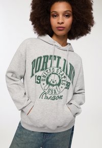 Felpa grigia in cotone con cappuccio, caratterizzata da testo grafico verde con la scritta "Portland", un'illustrazione di un orso e una tasca a marsupio.