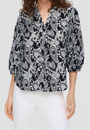 Frau trägt eine schwarze Bluse mit weißen Paisley- und Blumenmustern und weiße Hosen, mit dreiviertel Ärmeln, die an den Bündchen gerafft sind.