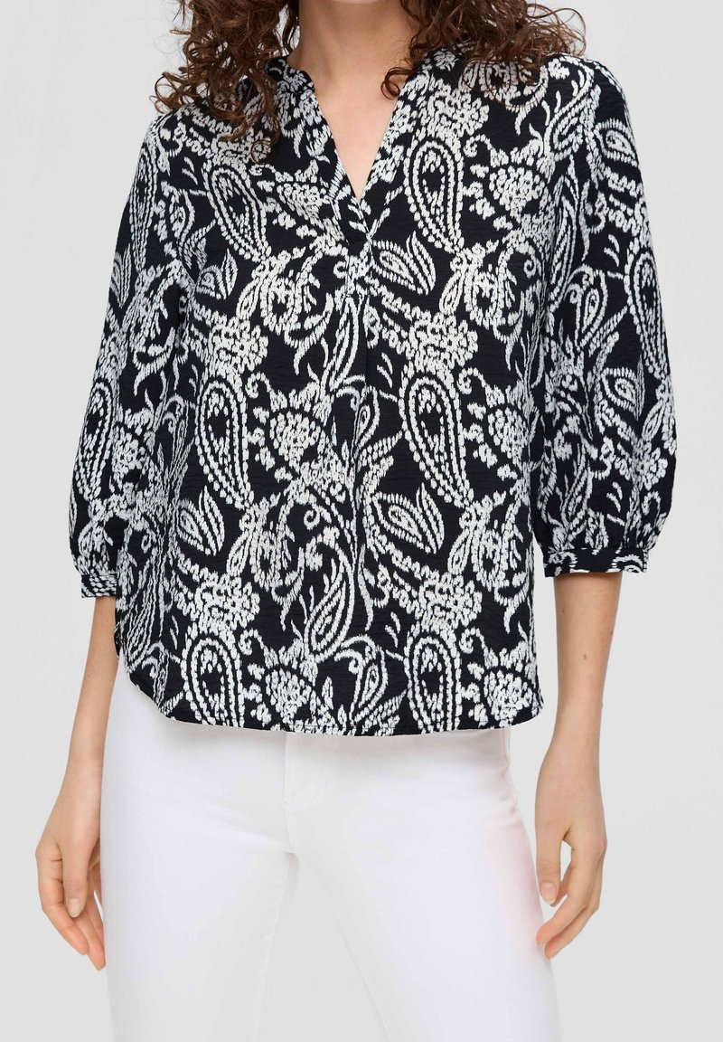 Femme portant un chemisier noir avec des motifs cachemire et floraux blancs et un pantalon blanc, avec des manches trois-quarts froncées aux poignets.