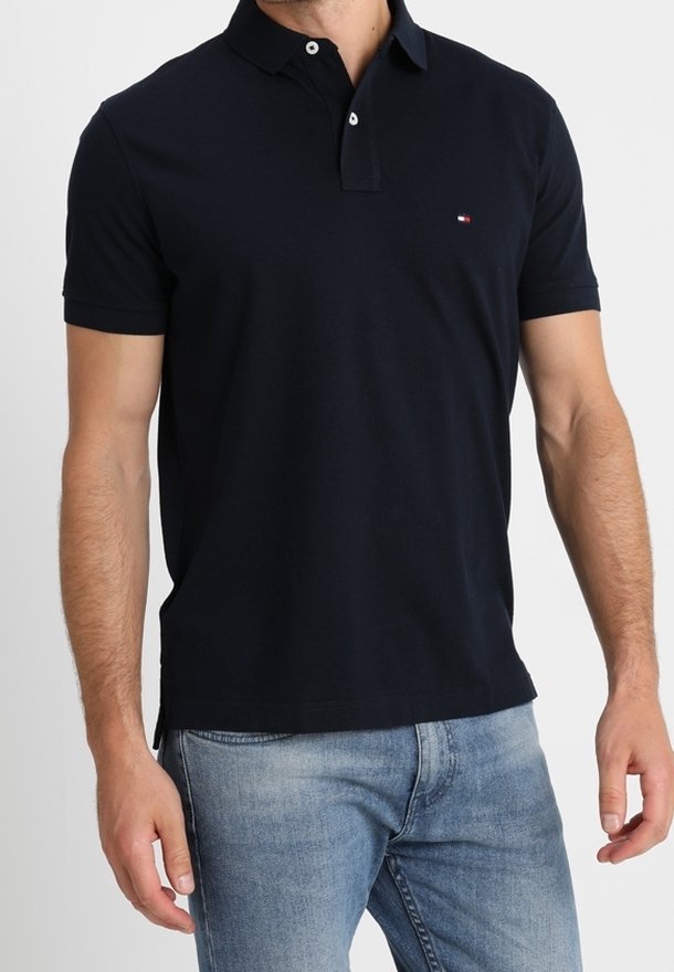 Homme portant un polo noir à manches courtes avec des boutons blancs et un jean bleu clair, debout devant un fond uni.
