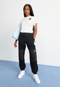 Tricou alb cu un mic logo negru, asociat cu pantaloni negri din fleece cu material texturat și un panou negru solid. Adidași albi.