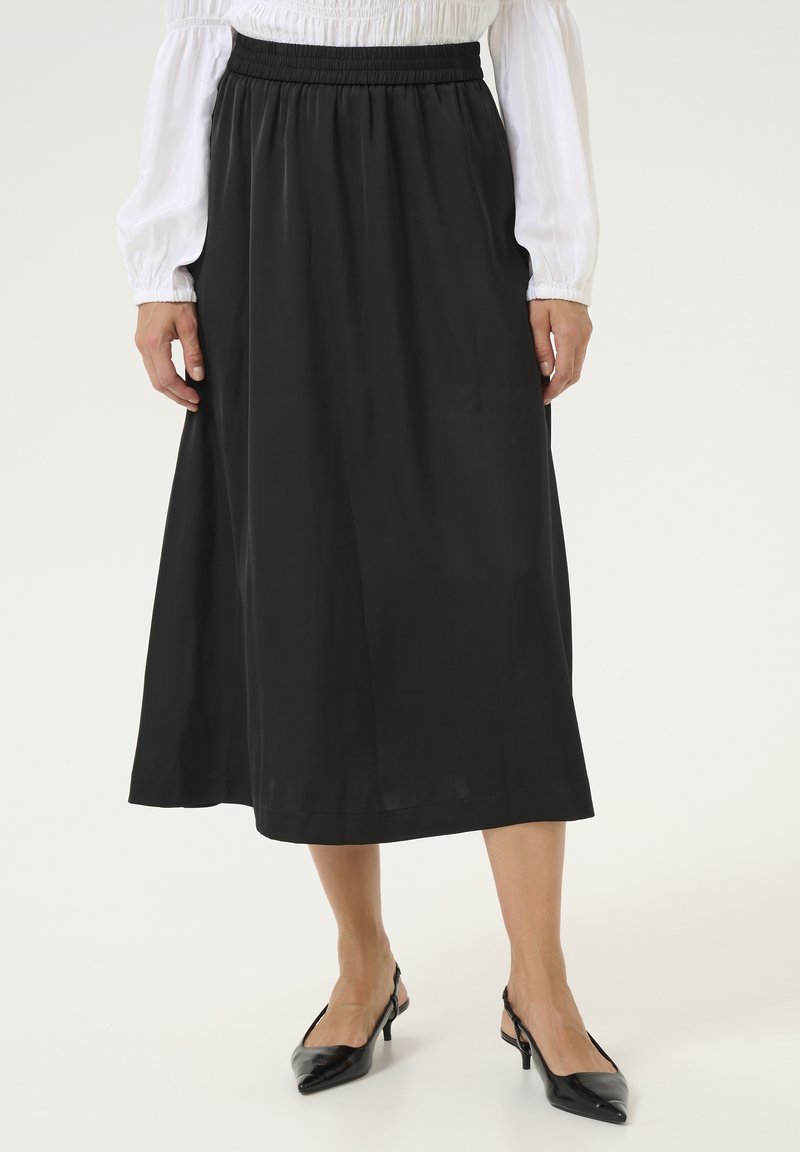 Ženska v črni midi krilu, beli bluzi z dolgimi rokavi in črnimi slingback čevlji s konico stoji pred belo ozadje.