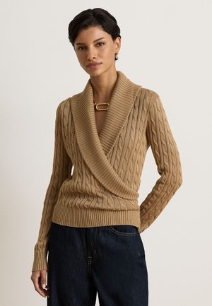 Femme portant un pull beige à torsades avec un grand col châle et un jean bleu foncé, debout devant un fond clair uni.