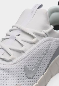 Κοντινό πλάνο λευκού αθλητικού παπουτσιού Nike που δείχνει ύφασμα από πλέγμα, ασημί λογότυπο Swoosh, μαλακή σόλα και λευκά κορδόνια σε λείο γλώσσα με ταινία τράβηγματος.